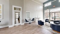 Located at 5005 Community Circle in Charlotte, NC, this thoughtfully designed single-floor space serves as a polished community gathering or leasing hub. The interior immediately impresses with dramatically soaring double-height ceilings that amplify the sense of openness throughout. Walls are finished in a refined palette of soft gray and deep teal accents, complemented by crisp white wainscoting and decorative panel molding that lend a sophisticated, contemporary character.

The main living area is anchored by light-toned wood-look flooring that runs seamlessly throughout, creating warmth and visual continuity. A striking marble-surround fireplace with a sleek linear insert serves as a bold focal point, paired with stylish navy barrel chairs arranged atop a textured area rug. Floor-to-ceiling glass entry doors flood the space with natural light and provide panoramic views of the exterior surroundings.

A dedicated workspace features a dark desk and seating, ideal for staff or administrative use. Distinctive circular pendant lighting adds a modern touch overhead. Decorative sculptural branches and minimal greenery contribute organic warmth. The connected hallway leads to additional areas, offering functional flow throughout the space. Together, these elements create an inviting, professional environment that balances comfort with contemporary design.