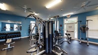 This fitness center offers a practical and functional workout environment designed to accommodate a range of exercise needs. The space is anchored by a large multi-station cable and weight machine, providing users with options for strength training across multiple muscle groups simultaneously. Additional cardio equipment, including a stationary bike, is positioned near a bright, glass-paneled door that allows natural light to fill the room and offers a view of the greenery outside. The bold blue accent walls give the space an energetic atmosphere, complemented by a dark rubber flooring that provides durability and traction underfoot. Overhead, fluorescent lighting ensures the entire area is well-lit, while ceiling fans promote air circulation throughout the room. A textured ceiling adds a subtle architectural detail to the space. The layout is thoughtfully arranged to allow users to move freely between equipment. Safety features, including a fire extinguisher and emergency exit signage, are clearly visible. The glass exit door leading outdoors enhances the sense of openness, making the fitness center feel inviting and well-ventilated for an effective workout experience.