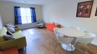 This single-floor space offers a welcoming and functional layout that seamlessly blends living and dining areas into one cohesive open-plan environment. The room is finished with warm hardwood-style flooring throughout, lending a clean and consistent look that ties the space together.

The living area is furnished with a comfortable sofa and a vibrant, multi-patterned accent chair positioned near a large window dressed with deep navy curtains, which allow for both light control and a bold pop of color. A sleek, clear acrylic coffee table adds a modern, airy touch to the seating arrangement. A vivid orange storage console serves as a striking focal point along the wall, providing both visual interest and practical storage.

The dining area features a contemporary white round table paired with curved, modern white chairs with chrome legs, creating a stylish and inviting setting for meals. Natural light filters into the space through the large window, brightening the neutral cream-toned walls and enhancing the overall sense of openness.

The space is characterized by its clean lines, minimalist approach, and thoughtful use of color accents, making it a practical yet visually engaging environment suitable for everyday living.