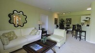 This single-floor space offers an open, airy layout that seamlessly connects the living and dining areas, creating a cohesive and functional flow. The living area is anchored by a plush white sofa adorned with patterned accent pillows, complemented by a striped armchair in soft green tones. A dark-framed coffee table with a woven surface serves as a stylish focal point, while a decorative quatrefoil mirror on the wall adds an elegant touch. Track lighting along the ceiling provides adjustable illumination, enhancing the warm, inviting atmosphere. The walls are painted in a calming light green hue, contributing to the bright and refreshing ambiance throughout. Lush potted plants are thoughtfully placed to bring natural life into the interior. The dining area, visible from the living space, features a dark wood table surrounded by classic black chairs, framed botanical artwork adorning the walls above. Light-toned carpeting runs throughout, adding warmth underfoot. The overall design balances casual comfort with refined detail, making the space feel welcoming and well-appointed.