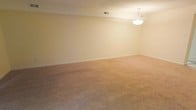 This two-story space offers a clean, understated layout designed for comfortable living. The image showcases a generously sized room featuring wall-to-wall beige carpeting that extends across the full floor area, providing a warm and soft underfoot experience. The walls are finished in a soft, creamy neutral tone that contributes to a bright and open atmosphere throughout the space.

A classic bowl-style pendant light fixture hangs from the ceiling, providing centralized overhead illumination and adding a simple, timeless aesthetic touch to the room. The ceiling features a standard textured finish, and a ceiling vent indicates a central HVAC system for climate control. White baseboards frame the perimeter of the room, offering a clean contrast against the neutral wall color and carpeted floor.

The room's generous proportions allow for flexible furniture arrangement, making it well-suited for use as a living or recreational area. The overall ambiance is calm and neutral, offering a blank canvas for personalization. As a two-floor property, additional living space, bedrooms, or functional areas are distributed across both levels.