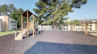 This outdoor playground area offers a well-equipped recreational space designed for active play. The ground is fully covered with dark wood chip mulch, providing a safe and impact-absorbing surface throughout the entire play area. A wooden play structure anchors one side of the space, featuring an elevated platform accessible via a staircase with railings, along with a slide, making it suitable for children of various ages. On the opposite side, a traditional swing set with multiple swings provides additional play options.

Several large, mature pine trees are positioned throughout the area, offering natural shade and contributing to a pleasant outdoor atmosphere on sunny days. The trees lend a sense of established greenery and visual depth to the space. The playground is situated adjacent to a multi-story residential building, visible in the background, suggesting this is a shared community amenity. The surrounding environment is open and spacious, allowing ample room for free play between the structured equipment. Overall, the area is functional, well-maintained, and provides a versatile outdoor setting for recreational use.