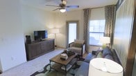 This single-floor residence centers around a comfortable and thoughtfully designed living room. The space is finished with light-toned walls that create a bright, neutral backdrop, complemented by plush carpeting underfoot. A ceiling fan with an integrated light fixture is mounted overhead, providing both air circulation and ambient lighting. Natural light fills the room through a large window dressed with patterned curtains, and a glazed door provides access to an outdoor area, further enhancing the sense of openness. The living room is laid out to accommodate a full seating arrangement, with space for a sofa, accent chair, and a rustic wood coffee table set atop a patterned area rug. A low-profile media console with cabinet storage anchors one wall, suitable for housing a flat-screen television and related media components. A tall floor lamp adds warm, layered lighting to the room. The overall ambiance is relaxed and functional, combining neutral tones with natural wood accents and green decorative touches to create an inviting, livable atmosphere.