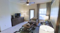 This single-floor residence centers around a comfortable and thoughtfully designed living room. The space is finished with light-toned walls that create a bright, neutral backdrop, complemented by plush carpeting underfoot. A ceiling fan with an integrated light fixture is mounted overhead, providing both air circulation and ambient lighting. Natural light fills the room through a large window dressed with patterned curtains, and a glazed door provides access to an outdoor area, further enhancing the sense of openness. The living room is laid out to accommodate a full seating arrangement, with space for a sofa, accent chair, and a rustic wood coffee table set atop a patterned area rug. A low-profile media console with cabinet storage anchors one wall, suitable for housing a flat-screen television and related media components. A tall floor lamp adds warm, layered lighting to the room. The overall ambiance is relaxed and functional, combining neutral tones with natural wood accents and green decorative touches to create an inviting, livable atmosphere.
