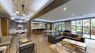 Located at 401 S Justison St in Wilmington, Delaware, this contemporary amenity lounge offers a thoughtfully designed communal living space with a seamless blend of style and function. The single-floor layout is anchored by a spacious open-plan living area furnished with plush sectional seating, a billiards table, and a geometric-patterned area rug that adds a vibrant touch underfoot. Wood-look flooring runs throughout, complementing the warm walnut wood wall panels that house a flat-screen television, creating a striking media focal point. Adjacent to the main lounge, a bar counter with high seating sits beneath a bold chevron-patterned accent wall flanked by decorative sconce lighting, ideal for social gatherings. Floor-to-ceiling glass walls on one side of the lounge flood the space with natural light and provide direct views of the outdoor pool and landscaped courtyard beyond. A welcoming lobby entrance features a dramatic multi-bulb chandelier, mid-century modern seating arrangements, and glass-panel doors that open to the exterior. A dedicated fitness room, visible through glass partitioning, offers residents convenient access to workout equipment. The overall ambiance is modern and inviting, with layered lighting, refined materials, and thoughtful design throughout.