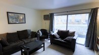 This single-floor space centers around a carpeted living room designed for comfortable everyday use. The room is furnished with dark upholstered seating, including a sofa, loveseat, and armchair, arranged around a rectangular coffee table. A large sliding glass door dominates one wall, dressed with dark curtain panels, and allows generous natural light to fill the room while offering a view of the surrounding outdoor area. A wall-mounted air conditioning unit is positioned near the window, providing in-unit climate control. The neutral-toned walls and light ceiling contribute to a clean, understated aesthetic. A framed piece of wall art adds a simple decorative touch. The carpeted flooring extends throughout the living area, lending a soft, casual feel to the space. Overall, the living room presents a functional and practical layout suited for relaxed, day-to-day living.