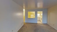 This single-floor space offers a straightforward, functional layout designed for practical everyday living. The living area is finished with wall-to-wall carpeting and painted in a neutral light blue-gray tone, creating a calm, understated atmosphere. Smooth flat ceilings and clean white trim provide a simple, no-frills aesthetic throughout.

A defining architectural feature is the pass-through window connecting the living area to the kitchen, framed with built-in shelving above. This opening enhances the sense of connectivity between the two spaces while maintaining a subtle visual separation. The pass-through also includes a small countertop ledge, useful as a serving or casual dining surface.

The kitchen is separately accessible and features white cabinetry along the walls, white appliances including a refrigerator and range, and light-toned flooring that contrasts with the carpeted living area. Additional countertop workspace and upper cabinets provide ample storage capacity. A secondary kitchen area or utility space is partially visible, suggesting further functional square footage beyond the main kitchen. Overall, the space is clean, vacant, and move-in ready.