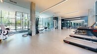 Located at 1250 Avenue Thérèse-Lavoie-Roux in Montréal, this contemporary fitness facility offers a generously sized, single-floor exercise room designed to support a full range of training activities. The space is defined by its striking floor-to-ceiling glass walls, which flood the interior with abundant natural light and provide views of the surrounding greenery, creating an energizing atmosphere for workouts.

The architectural design features exposed concrete columns and a clean, neutral palette of grey and teal tones, complemented by polished concrete flooring and sleek linear lighting overhead. The open layout is thoughtfully organized to accommodate multiple training zones without feeling crowded.

The cardio section is equipped with a row of treadmills and a rowing machine, while a dedicated strength training area includes Atlantis power racks, adjustable benches, and a comprehensive dumbbell rack with a full weight selection. Spinning bikes line one side of the room for cycling workouts, and functional training accessories such as TRX suspension systems and balance equipment are also on hand. Large mirrors along the walls further enhance the sense of space while allowing users to monitor their form. The facility strikes a balance between professional-grade equipment and a bright, welcoming environment suited to a wide variety of fitness routines.