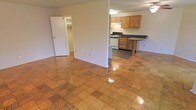 This single-floor space showcases a thoughtfully arranged open-concept layout that connects the living area to the kitchen, creating a sense of spaciousness throughout. The standout flooring feature is the polished parquet wood tile, which spans the main living area in a warm checkerboard pattern, lending character and a classic aesthetic to the space.

The kitchen is positioned at the rear and is equipped with oak cabinetry in a warm honey finish, dark countertops, and a white gas range. An extended countertop peninsula provides additional workspace or casual dining possibilities, and upper cabinets offer ample storage. A ceiling-mounted light fixture illuminates the kitchen area effectively.

A structural column divides the open living space, subtly defining zones without compromising the overall openness. Neutral, light-toned walls throughout the space provide a clean, versatile backdrop suitable for various interior styles.

A doorway visible from the main living area leads to an additional room featuring a walk-in closet, expanding the functional storage capacity of the unit. A ceiling fan is present, contributing to comfortable air circulation. Overall, the space presents a straightforward, functional layout with durable finishes.