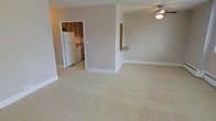 This single-floor residence offers a practical and comfortable layout centered around an open living area. The spacious main room features soft, neutral-toned wall-to-wall carpeting that creates a warm and inviting atmosphere, complemented by clean white baseboards and a ceiling fan with integrated lighting for year-round comfort. Neutral beige walls throughout contribute to a bright and airy feel. The kitchen is accessible via a doorway from the living area and is distinguished by easy-clean tile flooring, wood-toned cabinetry, and standard appliances including a full-size white refrigerator. A convenient pass-through counter connects the kitchen to the living space, making it ideal for casual dining or entertaining. The open-concept design between the living room and kitchen area maximizes the sense of space and encourages a natural flow throughout the home. The straightforward, functional layout makes efficient use of the available floor plan, providing a comfortable everyday living environment. Overall, the space presents a clean, move-in-ready interior with a cohesive neutral palette that offers a versatile backdrop for a variety of furnishing styles.