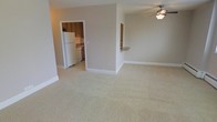 This single-floor residence offers a practical and comfortable layout centered around an open living area. The spacious main room features soft, neutral-toned wall-to-wall carpeting that creates a warm and inviting atmosphere, complemented by clean white baseboards and a ceiling fan with integrated lighting for year-round comfort. Neutral beige walls throughout contribute to a bright and airy feel. The kitchen is accessible via a doorway from the living area and is distinguished by easy-clean tile flooring, wood-toned cabinetry, and standard appliances including a full-size white refrigerator. A convenient pass-through counter connects the kitchen to the living space, making it ideal for casual dining or entertaining. The open-concept design between the living room and kitchen area maximizes the sense of space and encourages a natural flow throughout the home. The straightforward, functional layout makes efficient use of the available floor plan, providing a comfortable everyday living environment. Overall, the space presents a clean, move-in-ready interior with a cohesive neutral palette that offers a versatile backdrop for a variety of furnishing styles.
