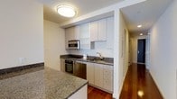 Unit 608 is a single-floor, one-bedroom, one-bathroom residence thoughtfully designed with a clean, contemporary aesthetic. The kitchen serves as a standout feature, showcasing sleek white cabinetry that extends to the ceiling for generous storage, complemented by dark granite countertops that provide both visual contrast and a durable workspace. Stainless steel appliances, including a built-in oven, microwave, and dishwasher, lend the kitchen a polished, modern feel. A double-basin sink sits beneath upper cabinetry, completing the functional layout. A kitchen island with matching granite adds additional prep and serving space. Rich hardwood flooring runs throughout the unit, adding warmth and continuity to the space. Flush-mount ceiling lighting provides bright, even illumination. A hallway connects the kitchen and living areas to the bedroom and bathroom, maintaining a logical and efficient flow throughout the residence. The overall ambiance is modern and well-appointed, with cohesive finishes and a neutral color palette that creates a welcoming and functional living environment.