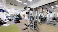 This expansive fitness center is thoughtfully organized into distinct training zones, offering a comprehensive workout environment for users of all fitness levels. The facility's architecture features high ceilings with recessed and flush-panel lighting throughout, arched transom windows, and large floor-to-ceiling windows that flood the space with natural light and provide views of an outdoor pool and tropical landscaping.

The cardio zone lines one side of the facility, featuring a full row of Life Fitness treadmills, StairMaster steppers, and stationary bikes, each positioned facing the large windows and equipped with individual monitors. Overhead, a mounted track of flat-screen televisions ensures entertainment throughout workouts.

The strength training area is generously proportioned, housing multiple Life Fitness and Hoist cable machines, a dual cable functional trainer, and a comprehensive rack of dumbbells along mirrored walls. Rubber sport flooring covers the free weights section, while neutral carpet tile defines the cable and machine areas.

A dedicated functional training zone features stacked tractor tires, kettlebells, medicine balls, and green turf matting, supporting agility and conditioning work. Large mirrors are positioned throughout, enhancing the sense of spaciousness. French doors connect sections of the facility, and ceiling fans contribute to overall comfort.