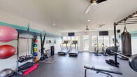 Located at 222 Kingfisher Way in Royal Palm Beach, Florida, this fitness center delivers a comprehensive workout facility within a single, open-plan space. The interior is finished with durable dark rubber flooring and bright white walls accented by a distinctive teal and navy horizontal stripe, lending the room a clean, energetic aesthetic.

The cardio zone features multiple treadmills, stationary cycling bikes, and an elliptical trainer, all positioned near large windows and glass French doors that fill the space with natural light and offer views of the surrounding outdoor area. Ceiling fans and a cassette-style air conditioning unit keep the environment comfortable throughout Florida's warm climate.

The strength and functional training area is equally well-appointed, housing a full power rack with a barbell and weight plates, a dumbbell rack, kettlebells, a cable machine, medicine ball storage, resistance bands, stability balls, a punching bag, and a flat adjustable bench. Wall-mounted televisions add to the amenity-forward atmosphere.

Recessed lighting complements the abundant natural light from the multiple windows, ensuring the space remains bright and inviting at all hours. The fitness center presents a versatile, fully outfitted environment suitable for a wide range of training styles and fitness goals.