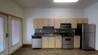 This single-floor unit offers a clean, functional layout designed for comfortable everyday living. The entryway opens through a corridor with warm wood-tone trim and durable hardwood-style plank flooring that flows consistently throughout the space, creating a cohesive and welcoming aesthetic.

The main living area is bright and airy, benefiting from generous natural light streaming in through large windows and a set of classic French doors that open to the exterior. The white walls and light-toned baseboards contribute to a fresh, neutral atmosphere that is easy to personalize.

The kitchen is efficiently designed and fully equipped with light maple cabinetry offering ample upper and lower storage. Dark countertops provide a practical contrast against the cabinetry, while the stainless steel appliances — including an electric range, over-the-range microwave, and dishwasher — lend a modern touch. A stainless steel sink is centrally positioned beneath the upper cabinets, making the workspace both organized and practical. The kitchen connects seamlessly to the rest of the living space, enhancing the open feel of the unit.

An electrical panel is conveniently accessible within the unit. Overall, the space presents a tidy, move-in-ready environment with a straightforward, modern design sensibility.