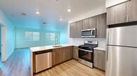 This contemporary single-floor unit offers a well-designed layout highlighted by light wood-style flooring that flows seamlessly throughout the space. The kitchen is a standout feature, equipped with a full suite of stainless steel appliances including a gas range, over-the-range microwave, refrigerator, and dishwasher. Cabinetry in a warm wood-grain finish provides generous storage, while white subway tile backsplash adds a clean, modern accent. A peninsula with a bold blue base and white quartz-style countertop serves as a functional focal point, offering additional prep space and a natural division between the kitchen and living areas. Recessed lighting illuminates the kitchen, complementing the natural light that filters through horizontal blinds. The living area is bright and open, with crisp white walls and multiple windows that invite an airy, welcoming atmosphere. A glass-panel door provides access to the exterior, adding additional light to the space. The bathroom features a full soaking tub with chrome fixtures, surrounded by large-format, stone-look tile in neutral tones that create a clean and relaxing environment. Overall, the unit presents a cohesive modern aesthetic with thoughtful finishes throughout.
