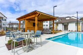 Located at 5905 Campbellton Rd SW in South Fulton, Georgia, this outdoor amenity area serves as a central gathering space within a contemporary residential community. The centerpiece is a generously sized swimming pool with striking blue tile accents and a wide concrete pool deck, offering ample space for lounging and recreation. Surrounding the pool, rows of chaise lounge chairs and patio seating with market umbrellas provide comfortable areas for relaxation.

A standout feature of the space is the timber-frame pavilion situated directly adjacent to the pool. Constructed with heavy wooden posts and a pitched roof, the structure houses an outdoor mounted television, upholstered seating, and a round fire table, creating an inviting outdoor living area suited for both daytime and evening use. Additional patio dining sets are positioned nearby for more casual gatherings.

The pool area is enclosed by black iron fencing, lending both security and a clean aesthetic. A well-appointed single-story clubhouse with large windows, dark trim, and a pitched roof borders the pool deck, adding architectural character to the setting. The surrounding multi-story residential buildings, featuring white siding and private balconies, frame the community's open-air environment cohesively.