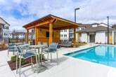 Located at 5905 Campbellton Rd SW in South Fulton, Georgia, this outdoor amenity area serves as a central gathering space within a contemporary residential community. The centerpiece is a generously sized swimming pool with striking blue tile accents and a wide concrete pool deck, offering ample space for lounging and recreation. Surrounding the pool, rows of chaise lounge chairs and patio seating with market umbrellas provide comfortable areas for relaxation.

A standout feature of the space is the timber-frame pavilion situated directly adjacent to the pool. Constructed with heavy wooden posts and a pitched roof, the structure houses an outdoor mounted television, upholstered seating, and a round fire table, creating an inviting outdoor living area suited for both daytime and evening use. Additional patio dining sets are positioned nearby for more casual gatherings.

The pool area is enclosed by black iron fencing, lending both security and a clean aesthetic. A well-appointed single-story clubhouse with large windows, dark trim, and a pitched roof borders the pool deck, adding architectural character to the setting. The surrounding multi-story residential buildings, featuring white siding and private balconies, frame the community's open-air environment cohesively.