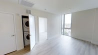 This contemporary two-floor space showcases a clean, minimalist design characterized by a crisp white interior palette and light-toned hardwood flooring throughout. The open layout is immediately inviting, benefiting from an abundance of natural light streaming through large floor-to-ceiling windows that frame expansive outdoor views of the surrounding cityscape, enhancing the sense of openness and connection to the environment.

The space is thoughtfully designed with practical conveniences built directly into the layout. A dedicated in-unit laundry closet houses a stacked washer and dryer, offering residents easy access without sacrificing living space. Ceiling-integrated HVAC vents and recessed lighting contribute to the modern, uncluttered aesthetic while ensuring comfort throughout.

The elevated position of the unit provides sweeping views, adding a premium quality to the living experience. Doorways connect seamlessly between rooms, maintaining a cohesive flow across the floor plan. The overall ambiance is bright, airy, and polished, reflecting a modern architectural sensibility that prioritizes both functionality and natural light throughout both levels of the home.