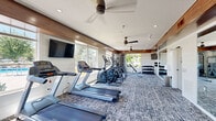Located in Shawnee, Kansas, this fitness center offers a well-appointed exercise room designed to support a comprehensive workout experience. The space features a thoughtful layout with distinct zones for both cardio and strength training. A row of treadmills and elliptical machines lines one side of the room, providing ample equipment for cardiovascular workouts, while a multi-station cable machine, adjustable weight bench, and a full dumbbell rack cater to strength training needs. Medicine balls and additional accessories round out the functional fitness offerings. Large windows wrap around much of the perimeter, flooding the space with natural light and offering views of the surrounding greenery and outdoor areas. Wood-trimmed tray ceiling details add a warm, modern aesthetic to the otherwise clean white shiplap walls. Multiple ceiling fans ensure comfortable air circulation throughout. Full-wall mirrors on one side visually expand the space and aid in proper form during exercise. Patterned carpet flooring provides cushioning underfoot, and wall-mounted flat-screen televisions offer entertainment during workouts. The room is accessible via multiple glass-paneled doors, reinforcing the bright and open character of this well-designed community fitness facility.