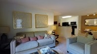 This single-floor residence offers a thoughtfully designed open-concept layout that connects the living area to the kitchen, creating an inviting and functional living environment. The living room is warmly decorated, featuring plush gray sofas accented with rose-toned throw pillows and a stylish coffee table that anchors the seating area. Two large gold-framed decorative wall panels add a touch of elegance to the neutral, softly lit space. A classic table lamp provides warm ambient lighting, complementing the room's cozy atmosphere. Carpeted flooring throughout the living area adds comfort underfoot. The space transitions naturally into the kitchen, which is separated by a partial wall and countertop peninsula. The kitchen is outfitted with black appliances, including a refrigerator, microwave, and range, set against crisp white cabinetry that keeps the space feeling bright and clean. Overhead lighting ensures the kitchen is well-illuminated for daily use. The open sightlines between rooms enhance the overall sense of space, making this residence feel connected and easy to navigate. The neutral color palette and understated décor create a welcoming ambiance throughout.