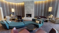 This refined lounge space presents an atmosphere of understated luxury through a carefully considered blend of architectural detail and interior design. The focal point of the room is a dramatic floor-to-ceiling stone fireplace clad in large-format marble-toned tiles, flanked by richly textured wave-patterned wall coverings that run the full height of the walls, adding depth and visual interest throughout the space. Cove lighting along the ceiling perimeter casts a warm, ambient glow that enhances the sophisticated palette of charcoal, taupe, and gold tones. The tray ceiling with recessed detailing adds architectural dimension and a sense of grandeur. Multiple seating vignettes are arranged throughout, including a curved teal sectional sofa as a central anchor, deep plum velvet loveseats, leather accent chairs, and dark upholstered club chairs — all complemented by brass-finished and stone-topped coffee tables. Wall-mounted sconces provide layered lighting and reinforce the upscale aesthetic. Sheer floor-to-ceiling drapery filters natural light from tall windows, softening the interior ambiance. A dining area with dark wood chairs occupies one corner, offering functional versatility. Rich patterned carpeting unifies the space, while corridor openings hint at additional areas beyond, completing this elegant and cohesive environment.