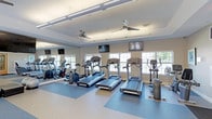 This fitness center offers a clean, well-organized workout environment designed to support a variety of exercise routines. The space is anchored by a row of treadmills and elliptical trainers positioned along a wall of large, multi-pane windows that invite generous amounts of natural light and provide views of the surrounding greenery outdoors. Full-length mirrors line one wall, visually expanding the room and allowing users to monitor their form during exercise. Overhead, ceiling fans are evenly distributed throughout, promoting comfortable air circulation during workouts. Wall-mounted flat-screen televisions are positioned at eye level for convenient viewing from the cardio equipment, enhancing the workout experience. The flooring consists of durable rubber athletic matting in a blue-toned palette, providing both safety and support underfoot. The ceiling features a recessed tray detail with integrated linear lighting that contributes to the bright, energizing atmosphere. The neutral gray walls and white trim create a crisp, modern aesthetic throughout. Overall, the fitness center is thoughtfully laid out to maximize usable floor space while maintaining an open, airy feel that encourages an active and motivating environment.