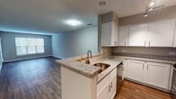 Located at 1710 Palmer St in Durham, NC, this single-floor unit presents a well-designed, open layout with consistent finishes throughout. The kitchen serves as a central feature, equipped with white shaker-style cabinetry, granite countertops with speckled detailing, and a peninsula that integrates a double-basin sink — ideal for both meal preparation and casual dining. Stainless steel appliances, including a refrigerator and electric range, complement the space, while track lighting above adds functional illumination. The kitchen opens directly into the living area, which is finished with warm wood-style flooring and neutral gray walls, creating a cohesive, airy feel. Natural light filters in through windows fitted with horizontal blinds. The bedrooms feature soft carpeting, neutral wall tones, and windows that bring in additional daylight. Closet space is available in the bedrooms, offering practical storage. The bathroom includes a full tub-shower combination with a curved curtain rod and wood-style flooring. Throughout the unit, clean white trim, textured ceilings, and a consistent neutral color palette contribute to a move-in-ready, contemporary ambiance.