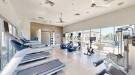 Located in Scottsdale, Arizona, this fitness center offers a well-equipped, single-floor workout space designed to support a comprehensive exercise routine. The room features a clean, modern aesthetic with neutral-toned walls, polished concrete flooring with large tile insets, and elevated ceilings fitted with ceiling fans for continuous air circulation.

The cardio section is prominently equipped with multiple treadmills, elliptical machines, and a stationary bike, each featuring individual digital display screens. Strength training options include a multi-station cable machine, a leg press/extension unit, a dumbbell rack, and an Octagon functional trainer system with suspension straps and accessories. A wall-mounted flat-screen television provides entertainment during workouts.

Natural light flows generously through expansive floor-to-ceiling windows and glass doors that line one side of the space, offering clear views of the outdoor amenity area, including a pergola and pool deck. This connection to the exterior creates an open, airy atmosphere that enhances the overall workout environment. The direct-access glass door provides convenient entry and exit to the outdoor area. The space is well-organized, clean, and purposefully laid out to maximize usability while maintaining an uncluttered, inviting atmosphere.