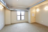 This single-floor unit offers a practical and efficiently designed layout that makes thoughtful use of its space. The living area is carpeted and open in character, featuring exposed ductwork along the ceiling that lends an industrial aesthetic. Wall-mounted sconce lighting provides a warm glow throughout, and a window allows natural light to filter into the space, offering a view of the surrounding building exterior.

The kitchen is compact yet well-equipped, outfitted with black appliances including a refrigerator, electric range, microwave, and dishwasher. Dark countertops contrast with light wood cabinetry, and open shelving above the sink area provides convenient storage. The kitchen transitions into the entryway, which features the same distinctive checkered tile flooring — a bold pattern that carries through into the bathroom.

The bathroom continues the checkered tile aesthetic and includes a walk-in shower with a clean white surround, a toilet, and a vanity with a dark countertop. Neutral gray walls throughout the unit create a cohesive, understated palette. Overall, the space presents a functional and straightforward design with an industrial edge, suited for those seeking a no-frills yet characterful living environment.