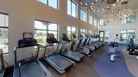 This contemporary two-story fitness facility is thoughtfully designed to provide a comprehensive workout experience across multiple specialized zones. The main level features a dedicated strength training area equipped with free weights, benches, squat racks, cable machines, and a variety of resistance equipment, all set against clean white walls and large windows that allow natural light to fill the space. A functional training studio offers ample open floor space alongside stability balls, resistance accessories, and wall-mounted equipment, with direct access to outdoor views through floor-to-ceiling windows. The cardio zone stands out architecturally with its soaring double-height ceilings, an expansive bank of large windows overlooking the surrounding landscape, and a striking geometric chandelier overhead — housing a full row of treadmills, ellipticals, and stair climbers. A central social and transition area on the lower level features a curved oval table with seating, a built-in display shelf, and warm accent walls in bold terracotta orange, creating a welcoming gathering point. A sleek open staircase with glass railings and a decorative perforated metal partition connects the two floors, complemented by elevator access. The upper level overlooks the main floor through curved glass balustrades, adding a sense of architectural continuity throughout the facility.