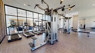Located at 355 W Burgundy St in Highlands Ranch, Colorado, this well-appointed fitness center offers a comprehensive workout environment across a single, open floor plan. The space is defined by its striking floor-to-ceiling black-framed glass wall, which floods the interior with natural light and provides views into an adjacent lounge or common area, creating a bright and energizing atmosphere.

The flooring throughout features a warm-toned herringbone pattern, lending a modern and polished aesthetic to the functional space. Ceilings are notably high, particularly in the free weights and functional training area, where large windows frame outdoor greenery and allow additional natural light to pour in.

Equipment is thoughtfully arranged to maximize usability. The cardio zone includes treadmills, ellipticals, stair climbers, a stationary bike, and a rowing machine. The strength training section is anchored by Cybex cable machines and multi-functional weight stations. A dedicated free weights area offers a full rack of dumbbells and a set of kettlebells displayed on a standing rack, complemented by a large wall mirror. Functional training equipment, including suspension trainers and vertical bar stations, occupies a spacious open zone.

The entry corridor features soaring ceilings, clean white walls, and herringbone flooring, providing a seamless and polished transition into the fitness space.