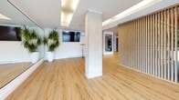 Located at 62-64 Boul de Lucerne in Gatineau, Quebec, this thoughtfully designed amenity facility combines modern aesthetics with functional fitness infrastructure across a single, generously sized floor. The entrance corridor sets an immediate tone of sophistication, featuring floor-to-ceiling wood-panel cabinetry with sleek black hardware, light hardwood flooring, and curated artwork adorning the walls.

The exercise room is a standout feature, designed to accommodate a comprehensive range of training activities. Durable rubber flooring defines the workout zones, while exposed ceilings painted in a warm terracotta hue lend an industrial character complemented by pendant lighting and structural concrete columns. The space is equipped with cardio machines including treadmills, stationary bikes, and ellipticals, as well as a full dumbbell rack and adjustable weight benches. Floor-to-ceiling mirrors amplify the sense of space, and a built-in wood storage wall adds warmth and organization.

The living room area offers a striking contrast, featuring light hardwood floors, exposed concrete columns, full-wall mirrors, vertical wood slat partitions, and an abundance of natural light from large windows. Lush potted plants and recessed cove lighting create a calm, welcoming ambiance. A wall-mounted television and open layout make this a highly adaptable communal space.