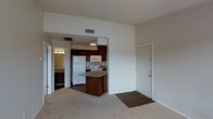 This single-floor residence offers an open-concept layout that seamlessly connects the living area and kitchen, creating a cohesive and functional living space. The carpeted living area provides a comfortable, neutral foundation that transitions into hard flooring at the entryway, marked by a classic paneled door.

The kitchen is outfitted with rich, dark wood cabinetry that provides ample storage, complemented by light-toned countertops. A peninsula extends from the main cabinetry, offering additional counter space and serving as a natural divider between the kitchen and living area. White appliances, including a refrigerator, electric range, and over-the-range microwave, are neatly integrated into the layout.

Ceiling vents are positioned throughout, ensuring consistent climate control across the space. An adjacent room, visible through a doorway, features a light fixture and additional cabinetry with countertop space, suggesting a utility or bathroom area.

Overall, the space maintains a clean, neutral color palette with beige walls and textured ceilings, contributing to a straightforward and practical living environment well-suited for everyday use.