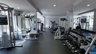 This fitness center offers a thoughtfully designed workout environment finished in a sleek, modern aesthetic with neutral gray tones throughout. The space features dark rubberized flooring that provides both durability and comfort underfoot, making it well-suited for a variety of exercise activities.

The room is outfitted with a thorough selection of equipment to support diverse fitness routines. Cardio machines, including treadmills and ellipticals, are lined along one side of the space, while multi-station cable and strength training machines are positioned to allow for efficient use of the floor plan. A dedicated free weights area features a full rack of dumbbells in graduating sizes, accompanied by a flat utility bench for targeted exercises.

Floor-to-ceiling mirrors are installed along the walls, enhancing the sense of openness and allowing users to monitor their form during workouts. Recessed lighting in the ceiling provides consistent, even illumination throughout the space. The layout is open and uncluttered, allowing for easy movement between stations. A clearly marked exit and clean white walls contribute to a safe and organized environment. Overall, this fitness center is a functional and well-appointed space designed to accommodate a broad range of fitness needs.