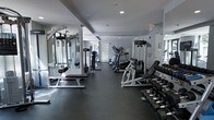 This fitness center offers a thoughtfully designed workout environment finished in a sleek, modern aesthetic with neutral gray tones throughout. The space features dark rubberized flooring that provides both durability and comfort underfoot, making it well-suited for a variety of exercise activities.

The room is outfitted with a thorough selection of equipment to support diverse fitness routines. Cardio machines, including treadmills and ellipticals, are lined along one side of the space, while multi-station cable and strength training machines are positioned to allow for efficient use of the floor plan. A dedicated free weights area features a full rack of dumbbells in graduating sizes, accompanied by a flat utility bench for targeted exercises.

Floor-to-ceiling mirrors are installed along the walls, enhancing the sense of openness and allowing users to monitor their form during workouts. Recessed lighting in the ceiling provides consistent, even illumination throughout the space. The layout is open and uncluttered, allowing for easy movement between stations. A clearly marked exit and clean white walls contribute to a safe and organized environment. Overall, this fitness center is a functional and well-appointed space designed to accommodate a broad range of fitness needs.