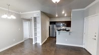 This well-appointed single-floor residence offers a thoughtfully designed open-concept layout that seamlessly connects the living and kitchen areas. The space is finished with rich, dark hardwood-style flooring that contrasts elegantly with the soft gray walls and crisp white trim, creating a clean and contemporary aesthetic throughout.

The kitchen is a standout feature, outfitted with dark espresso cabinetry, stainless steel appliances including a refrigerator, microwave, and range, and a peninsula countertop with an overhang that provides casual bar-style seating. A stylish pendant light fixture hangs above, adding a warm focal point to the culinary space.

Adjacent to the kitchen, a built-in open shelving unit with multiple tiers offers both functional storage and a decorative display area, adding architectural character to the open floor plan. The layout flows naturally into the living area, which benefits from recessed lighting that keeps the space bright and welcoming.

A hallway visible from the main living area leads to additional rooms, with white panel doors and consistent neutral tones maintaining a cohesive design language throughout. The vaulted ceiling detail above the kitchen adds a sense of openness and vertical dimension. Overall, the space presents a modern, functional, and inviting environment suited for comfortable everyday living.
