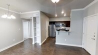 This well-appointed single-floor residence offers a thoughtfully designed open-concept layout that seamlessly connects the living and kitchen areas. The space is finished with rich, dark hardwood-style flooring that contrasts elegantly with the soft gray walls and crisp white trim, creating a clean and contemporary aesthetic throughout.

The kitchen is a standout feature, outfitted with dark espresso cabinetry, stainless steel appliances including a refrigerator, microwave, and range, and a peninsula countertop with an overhang that provides casual bar-style seating. A stylish pendant light fixture hangs above, adding a warm focal point to the culinary space.

Adjacent to the kitchen, a built-in open shelving unit with multiple tiers offers both functional storage and a decorative display area, adding architectural character to the open floor plan. The layout flows naturally into the living area, which benefits from recessed lighting that keeps the space bright and welcoming.

A hallway visible from the main living area leads to additional rooms, with white panel doors and consistent neutral tones maintaining a cohesive design language throughout. The vaulted ceiling detail above the kitchen adds a sense of openness and vertical dimension. Overall, the space presents a modern, functional, and inviting environment suited for comfortable everyday living.