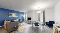 This single-floor residence offers a thoughtfully designed layout with a seamless blend of comfort and functionality. The open-concept living and dining area serves as the heart of the home, featuring light wood-style flooring, multiple windows dressed with grommet-top curtains that invite ample natural light, and a bold blue accent wall that adds a striking focal point to the living space. A modern pendant light fixture anchors the dining area, which flows naturally into the living room. The kitchen is equipped with two-tone cabinetry — white upper cabinets and gray lower cabinets — paired with white countertops, a classic subway tile backsplash, and black appliances including a range and built-in microwave. The bedrooms offer carpeted flooring, generous closet space with mirrored sliding doors, and large windows, with one bedroom featuring a distinctive chevron-patterned accent wall. Two bathrooms include warm wood-toned vanities, tile flooring, and well-lit mirror-and-vanity setups, with one finished in a rich teal accent color. A dedicated in-unit laundry area houses a full-size front-loading washer and dryer with overhead shelving for added storage, completing this practical and stylish residence.