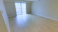 This single-floor living space features a clean, open layout designed to maximize comfort and functionality. The main living area is finished with smooth, light-toned hardwood flooring that reflects natural light beautifully, contributing to a bright and airy atmosphere throughout. Neutral beige walls provide a versatile backdrop, complementing a variety of interior styles and furnishings.

A prominent sliding glass door fitted with vertical blinds serves as the focal point of the room, allowing abundant natural light to flood the space while offering direct access to an outdoor area. The blinds provide flexible light and privacy control, making the space adaptable to different times of day.

Practical details include ceiling-mounted HVAC vents for climate control and crisp white baseboards that frame the room neatly. The open floor plan offers considerable flexibility in furniture arrangement, lending itself well to a variety of living arrangements. The overall ambiance is clean, bright, and move-in ready, with a simple, modern aesthetic that provides a welcoming and functional everyday living environment.