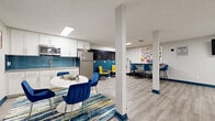 Located at 914 W Lake St in Fort Collins, Colorado, this thoughtfully designed single-floor space offers a dynamic, open-plan layout ideal for collaboration, productivity, and community gathering. The interior is defined by a clean, modern aesthetic with light gray luxury vinyl plank flooring, crisp white walls, and bold blue and yellow accents that bring energy and cohesion throughout.

A dedicated computer workstation area lines one wall, featuring a row of desks equipped with desktop computers, vibrant yellow ergonomic chairs, and decorative column dividers. A wall-mounted flat-screen TV completes this functional zone. Recessed and flush panel lighting ensures the space is well-lit throughout.

The social lounge area centers around a tall communal table with elegant blue upholstered bar chairs, complemented by a colorful piece of wall art and a noted complimentary Wi-Fi area. Nearby, a cozy dining nook with a round white table and blue chairs sits atop a multicolored area rug, adding warmth to the space.

A kitchenette with white cabinetry and a full-size stainless steel refrigerator provides practical amenities. Structural columns add architectural character while defining the open zones. A bold teal-painted entry door serves as a welcoming focal point, tying together the space's lively and inclusive atmosphere.