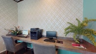 Located at 2120 Village Park Rd in Plant City, Florida, this single-floor business center is thoughtfully designed to provide a comfortable and functional environment for residents or visitors. The space features a dedicated computer workstation area outfitted with multiple desktop computers, keyboards, and a multifunction printer, all arranged along a built-in curved countertop. The workstation wall is adorned with an elegant geometric patterned wallcovering in neutral tones, adding a refined aesthetic to the functional setup. Upholstered gray chairs provide comfortable seating at each workstation. Complementing the work area is a casual lounge and meeting nook, highlighted by rich teal-painted walls and dark hardwood-style flooring. A round high-top table surrounded by a mix of white and gray upholstered chairs offers an inviting space for informal gatherings or collaborative work. A framed world map serves as a focal point on the accent wall, adding a sophisticated decorative touch. The room benefits from generous natural light streaming through large windows and French doors fitted with white horizontal blinds. Lush potted greenery and floral arrangements are placed throughout the space, contributing to a warm and welcoming atmosphere.