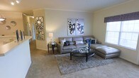 This single-floor residence features a spacious living area designed with a cohesive, contemporary aesthetic. The main living room is anchored by a large gray leather sectional sofa accented with decorative throw pillows in navy and silver tones, paired with a sleek oval glass coffee table resting atop a plush area rug. Crown molding along the ceiling adds a refined architectural detail, complementing the warm, neutral wall tones that lend the space a calm and welcoming ambiance. A decorative mirror with a geometric frame adorns one wall, enhancing the sense of depth and light in the room. Two bold abstract canvas prints in black, white, and blue serve as a striking focal point above the sofa, adding artistic character. Natural light filters in through a window dressed with a patterned valance, brightening the carpeted floors. An open sightline toward an adjacent area suggests a connected, flowing floor plan. The overall atmosphere is polished and inviting, combining modern design elements with comfortable living spaces suited for everyday use.