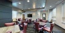This well-appointed single-floor space is designed to accommodate both formal gatherings and casual dining, combining functionality with refined aesthetics. The main dining area features multiple square tables surrounded by rich burgundy leather chairs, all set atop a striking dark carpet with sweeping white linear patterns that lend dynamic movement to the room. Recessed lighting illuminates the space evenly, while a wall-mounted flat-screen television and natural light from blinds-covered windows add practical amenities.

A semi-private meeting area is separated by elegant wood-framed glass doors with decorative art-glass panels, offering a more intimate setting with its own table and seating. The patterned carpet flows seamlessly throughout both areas, reinforcing visual continuity.

Along one wall, a built-in kitchenette serves the space with granite countertops, an under-counter refrigerator, and a warm pendant light overhead. Nearby, floating glass shelves display decorative vases and objects, adding a curated, gallery-like touch. Textured wall panels in muted taupe tones and layered lighting throughout contribute to a polished, professional ambiance suited for corporate or social events.