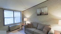 This single-floor space includes a living room that serves as a central gathering area, characterized by its neutral color palette and understated design. The walls feature a smooth textured finish in warm beige tones, lending a calm and cohesive atmosphere to the room. Soft wall-to-wall carpeting underfoot adds comfort and warmth throughout the space.

A large window with vertical blinds allows natural light to filter into the room, offering an outdoor view of surrounding trees and providing a connection to the outside environment. A baseboard heating unit sits beneath the window, ensuring year-round comfort. The ceiling features a subtle textured finish, adding modest visual interest overhead.

The living area is well-suited for relaxed everyday use, with enough room to accommodate a sofa and side furniture comfortably. Accent lighting from a table lamp positioned on a wooden end table contributes to a warm, inviting ambiance during evening hours. A framed cityscape artwork displayed on the wall adds a decorative focal point, complementing the room's muted tones. Overall, the space presents a functional and quietly stylish environment.
