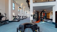 This expansive fitness center is designed to impress, combining striking architectural elements with a full suite of professional-grade exercise equipment. The space is defined by its dramatic double-height ceilings, which are lined with oversized windows that flood the interior with natural light and offer views of the surrounding greenery. Soft blue-grey carpet tiles extend throughout, complemented by crisp white walls accented with teal base molding for a modern, cohesive aesthetic.

The main workout area is thoughtfully organized into distinct zones. A dedicated cardio section houses a row of treadmills and elliptical machines, each positioned to take advantage of the expansive window views. Nearby, a functional training zone features a tire flip sled, suspension training rigs with resistance straps, and a multi-functional cable machine. A strength training area includes a variety of weight machines, benches, and a full dumbbell rack accompanied by a wall-mounted display screen and mirrored wall.

The entryway features a bold, large-scale abstract mural that serves as a vibrant focal point, alongside a water fountain station. A mezzanine railing is visible overhead, adding architectural depth. The overall ambiance is energetic yet polished, making this a well-equipped and visually engaging fitness facility.