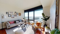 This well-appointed single-floor residence is defined by its dramatic floor-to-ceiling windows, which frame expansive views of a river or lake surrounded by lush treetops—visible from both the living room and the primary bedroom. The open living area features rich hardwood flooring, a curated gallery wall, and an abundance of natural light that streams in throughout the day, creating a bright and welcoming atmosphere.

The kitchen is outfitted with warm walnut cabinetry, stainless steel appliances including a gas range with a stainless hood, and light countertops. A central island with a butcher block top provides additional prep space and casual seating, making the kitchen both functional and visually appealing.

The primary bedroom continues the floor-to-ceiling window theme, offering the same stunning water views and generous natural light. It also features a compact closet system for organized storage. A second bedroom is carpeted and includes a walk-in closet, providing comfortable and practical accommodations.

The bathroom is clean and functional, featuring a wood-toned vanity with an undermount sink, a soaking tub and shower combination, and recessed lighting. Throughout the residence, the design balances modern finishes with warm, natural materials.