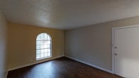 This single-story space in Arlington offers a clean, straightforward layout with notable architectural and design details. The main living area showcases rich, dark hardwood flooring that contrasts with crisp white baseboards running along the neutral-toned walls. One wall features a warm yellow accent, adding a touch of color to the otherwise understated palette.

A standout architectural feature is the arched window, which draws the eye and floods the room with natural light from the outdoors, where mature trees are visible beyond the glass. The window's classic grid-pane design adds character to the space. The textured popcorn ceiling adds a traditional element consistent with the overall aesthetic of the interior.

The open, unobstructed floor plan provides flexibility for various furniture arrangements and uses. Neutral gray walls on the opposing side of the room offer a versatile backdrop. Overall, the space presents a functional and well-lit interior with a blend of traditional design elements, including the arched window detail, hardwood-style flooring, and classic white trim work throughout.
