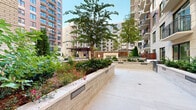 This expansive outdoor courtyard is a thoughtfully designed amenity space nestled within a modern multi-story residential complex. The courtyard is framed by contemporary brick and stone façades with balconied residences rising on all sides, creating a sense of enclosure while remaining open to natural light and sky.

At the heart of the space is a generous landscaped lawn, finished with manicured turf and set up for lawn games such as cornhole, making it an inviting area for casual outdoor recreation. Stone-clad raised planters filled with mature trees, columnar evergreens, flowering shrubs, and lush ground cover add depth and natural beauty throughout.

A standout feature is the elegant covered pavilion, constructed with stone columns and a rich wood-paneled ceiling. It houses a wall-mounted television and serves as a central gathering hub. Adjacent to the pavilion is a dramatic outdoor fireplace framed in polished marble, surrounded by wicker lounge chairs and accent tables — ideal for relaxed social gatherings.

Multiple outdoor dining areas are equipped with grills and dining sets, catering to residents who enjoy al fresco entertaining. Additional seating is scattered throughout, including Adirondack chairs and built-in benches. Wide stone-paved pathways connect each zone seamlessly. The overall ambiance is refined yet welcoming, blending upscale finishes with functional, community-oriented outdoor living.