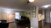 This single-floor space presents a functional, open layout that blends work and living areas seamlessly. The main area centers around a dark wood desk equipped with multiple computer monitors, making it suitable for office or administrative use. Adjacent to the workstation, a small dining table with dark wood chairs provides a casual seating area. The flooring throughout is dark hardwood, lending warmth and contrast to the light grey walls. A vibrant green accent wall serves as a backdrop to the workstation, adding a pop of color and visual interest to the space. Two framed city-themed art prints are displayed on a partial wall divider, contributing to an urban aesthetic. Natural light enters through a window along the far wall, brightening the space and offering a view of the greenery outside. A ceiling-mounted light fixture provides additional overhead illumination. A textured ceiling adds subtle architectural character. To one side, a partial view of a kitchen area is visible, featuring light wood cabinetry and what appears to be white appliances, suggesting convenient access to kitchen amenities. Overall, the space has a practical, urban-inspired character suited for both productivity and everyday use.
